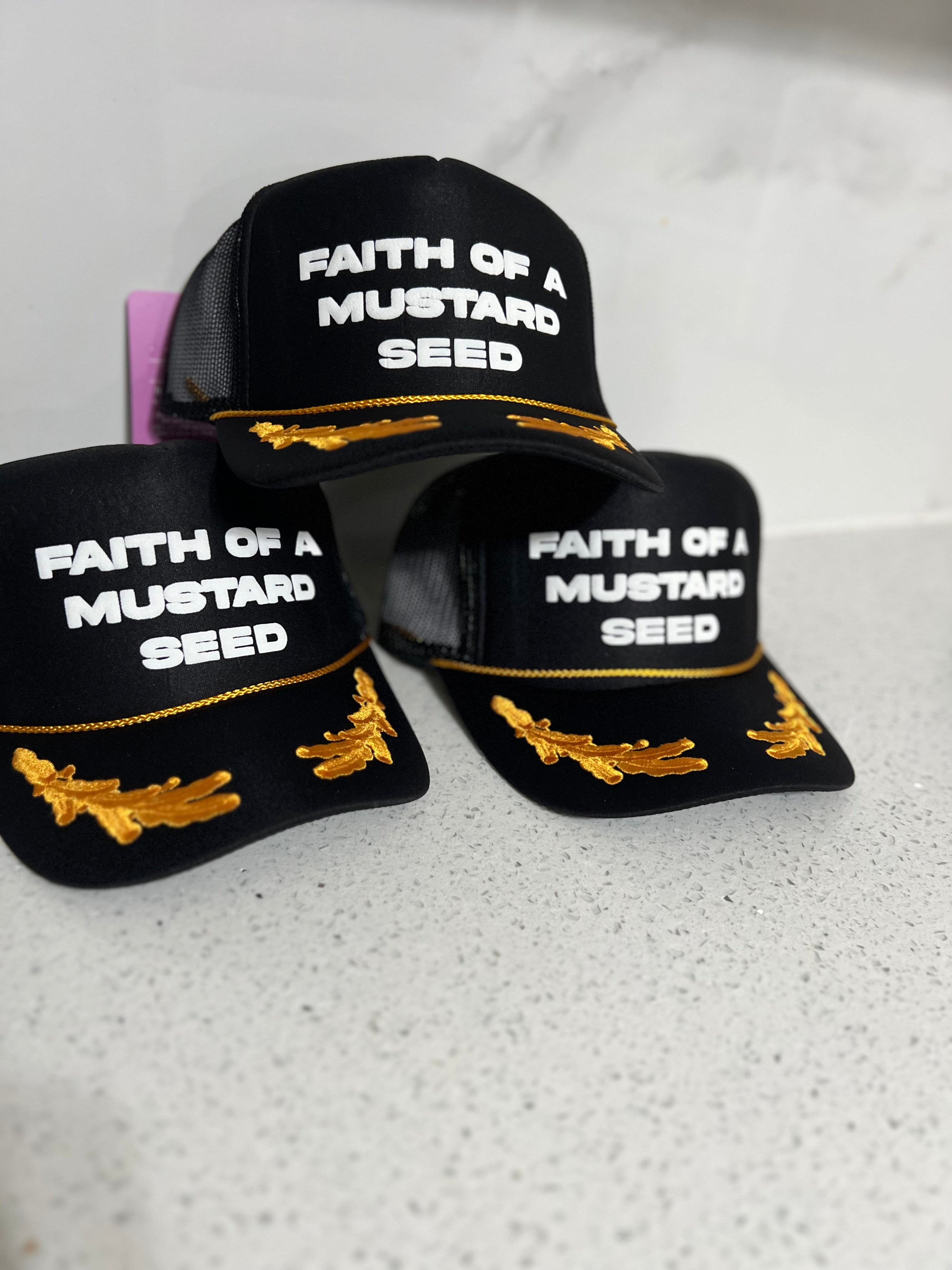 Black hats with 'Faith of a Mustard Seed' text and gold accents on a white surface